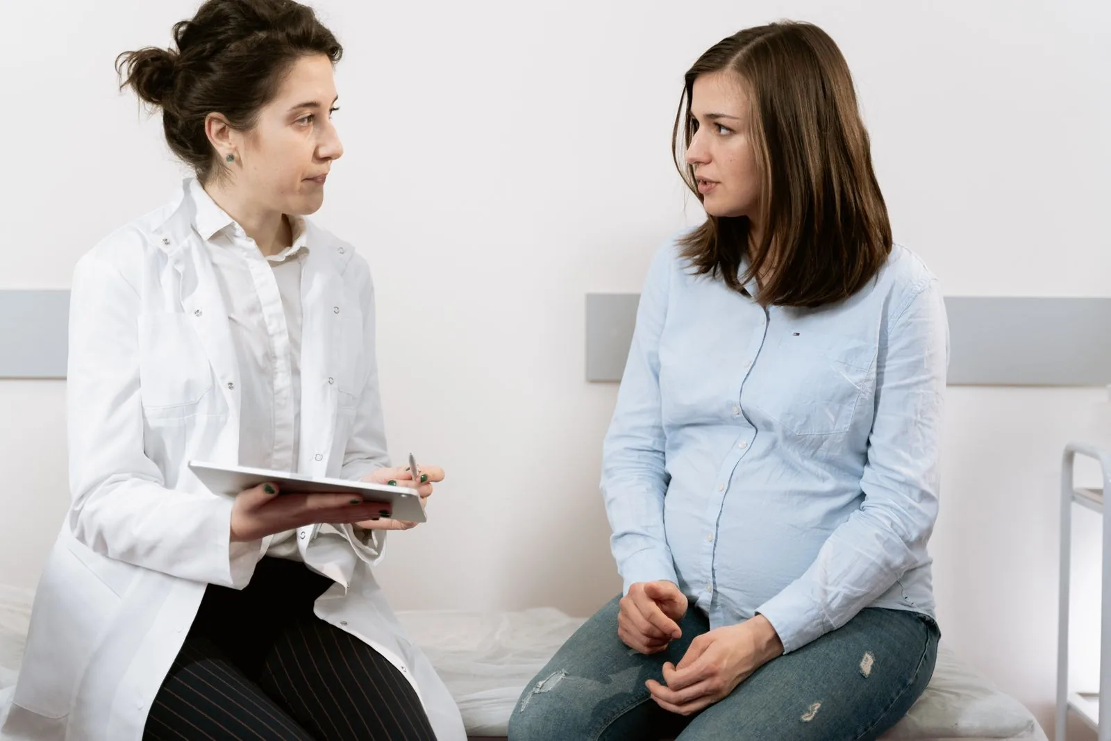 Pregnant woman sitting on an exam table in conversation with a clinician holding a chart
