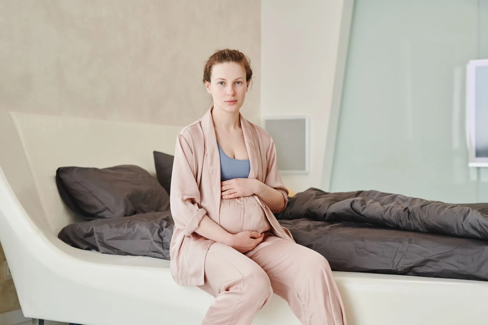 Pregnant woman in a blush robe sitting on the edge of a bed with both hands resting low on her bump